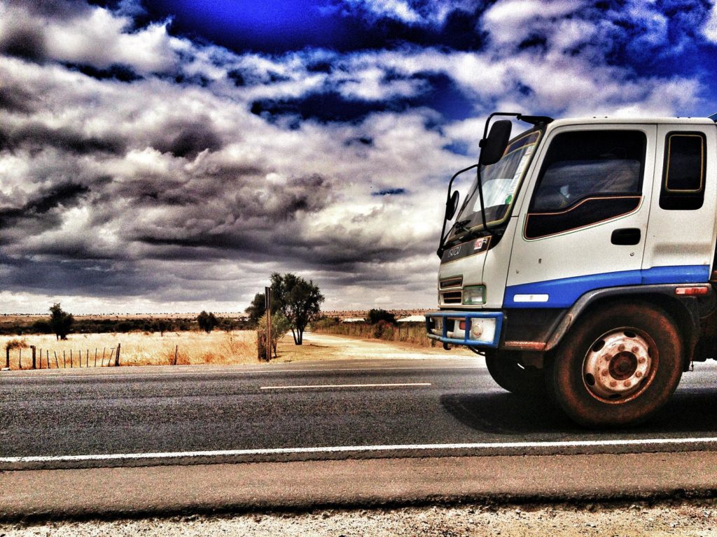 Na zdjęciu widać fragment ciężarówki, która jedzie po drodze. W tle jest mocno niebieskie niebo z dużą ilością białych chmur.