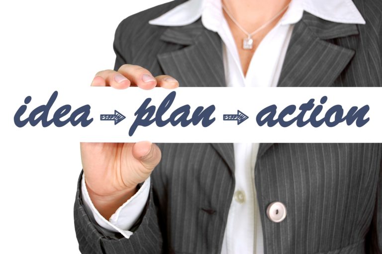 Na zdjęciu znajduje się człowiek(kobieta) ubrana w szarą marynarkę i białą koszulę, która trzyma w dłoni biały napis: Idea - Plan- Action.