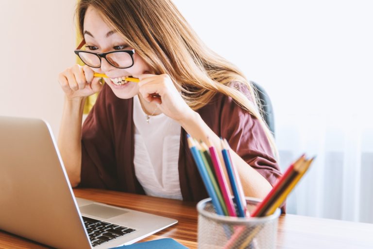 Na zdjęciu przy biurku siedzi młoda kobieta o długich włosach i w okularach. W ustach trzyma żółty ołówek. Siedzi przy biurku drewnianym, na którym stoi laptop i podajnik z kolorowymi ołówkami.