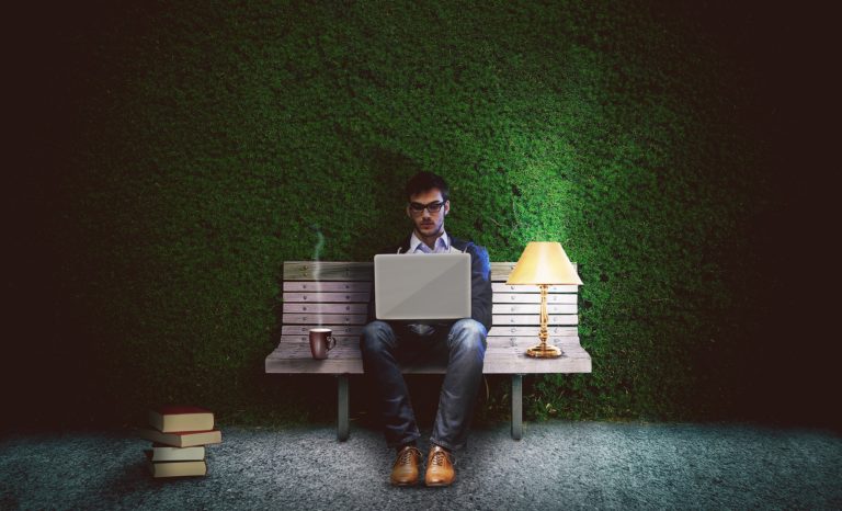 Na zdjęciu znajduje się młody mężczyzna siedzący na ławce w zielonym pokoju. Trzyma na kolanach laptopa, na którym pracuje.