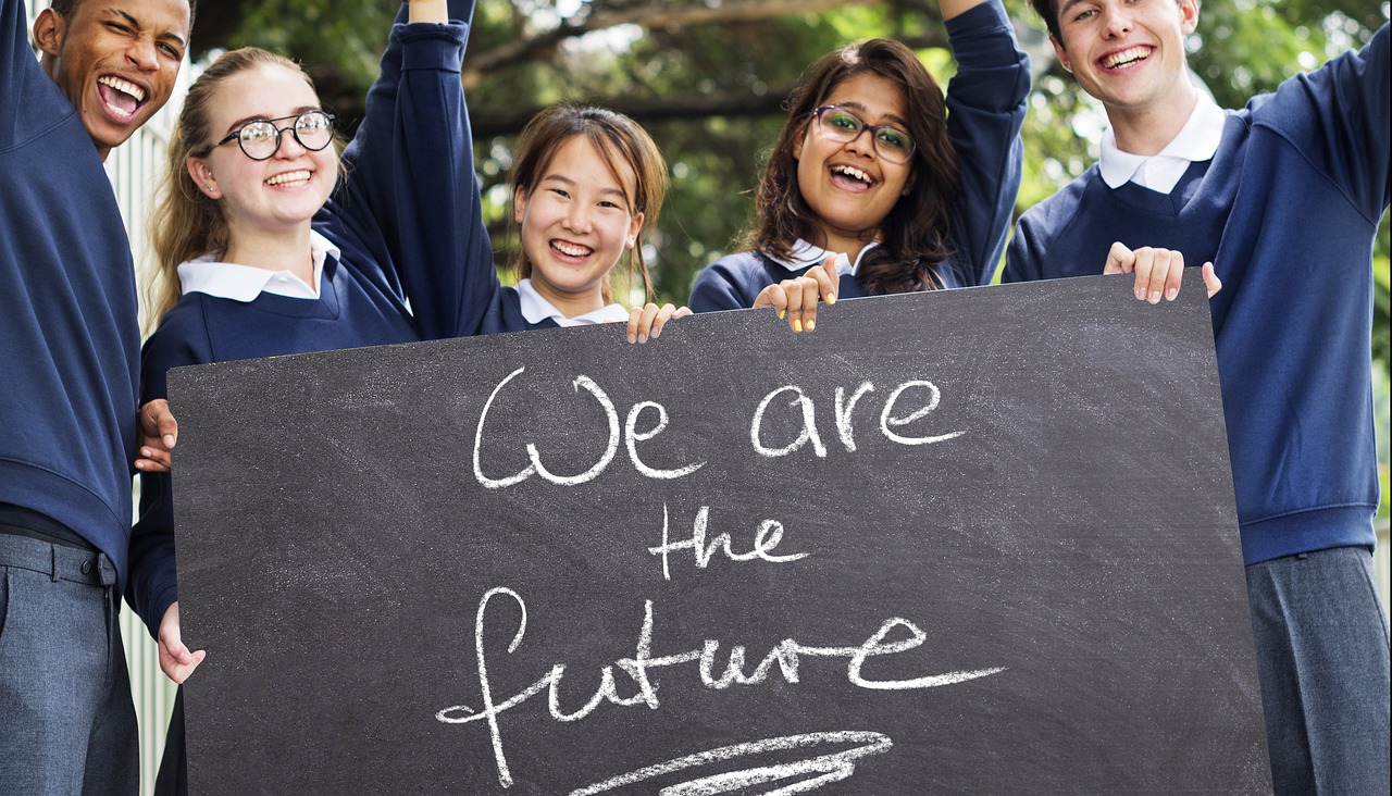 Na zdjęciu znajduje się grupka młodzieży - uczniów, którzy ubrani są w szkolne niebiesko-białe mundurki i trzymają w rękach czarną tablicę z napisem "We are the future"