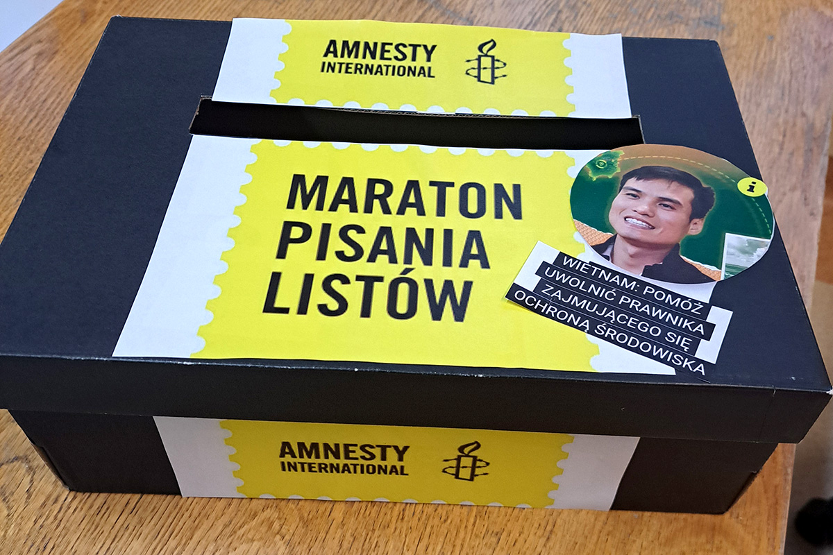 Na zdjęciu znajduje się kartonik z naklejką Maratonu pisania listów Amnesty International. Napisy są w kolorze czarnym na żółtym tle. Kartonik służy wkładaniu tam listów dla osób, które je napiszą.