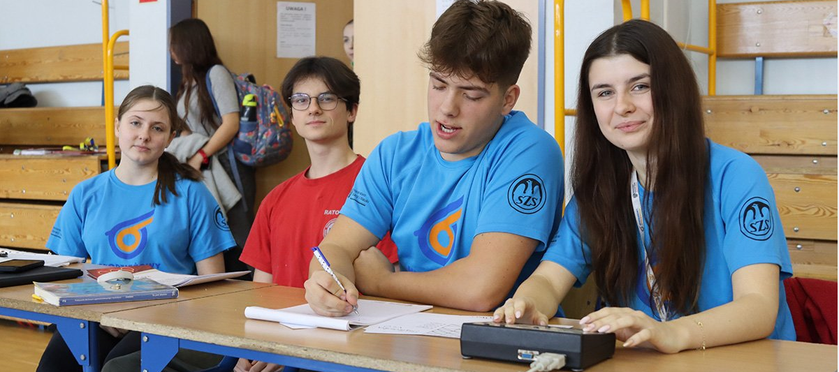 Na zdjęciu znajdują się uczniowie jednej ze szkół w Gdyni. Ubrani są w niebieskie i czerwone koszulki, siedzą przy biurkach i robią notatki.