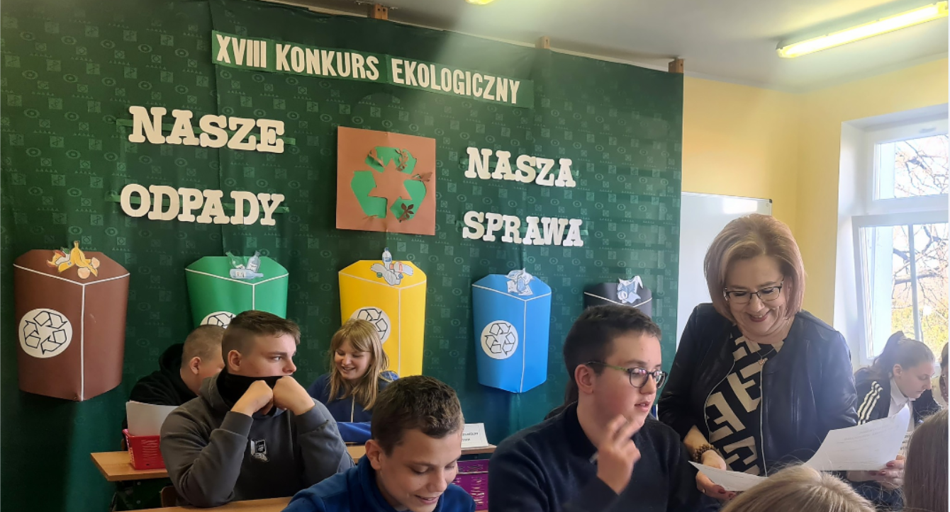 Na zdjęciu znajdują się uczniowie wraz z nauczycielem. Siedzą w ławkach szkolnych, a z tyłu na ścianie na zielonym tle wisi napis: Konkurs Ekologiczny pod nazwą" Nasze odpady, nasza sprawa.