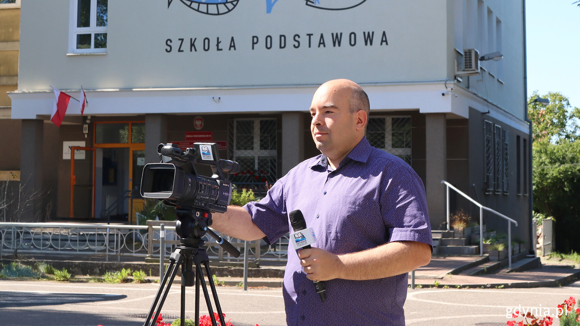 Mężczyzna trzymający mikrofon w lewej ręce ustawia parametry kamery, która stoi przed nim na statywie, w tle wejście i fasada budynki ZSO nr 6 w Gdyni